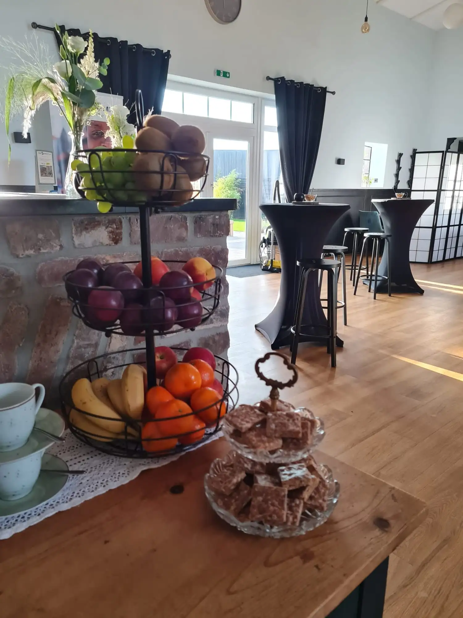 Etagere mit Obst und Gebäck auf dem Holztresen, dahinter eingedeckte Stehtische im Saal