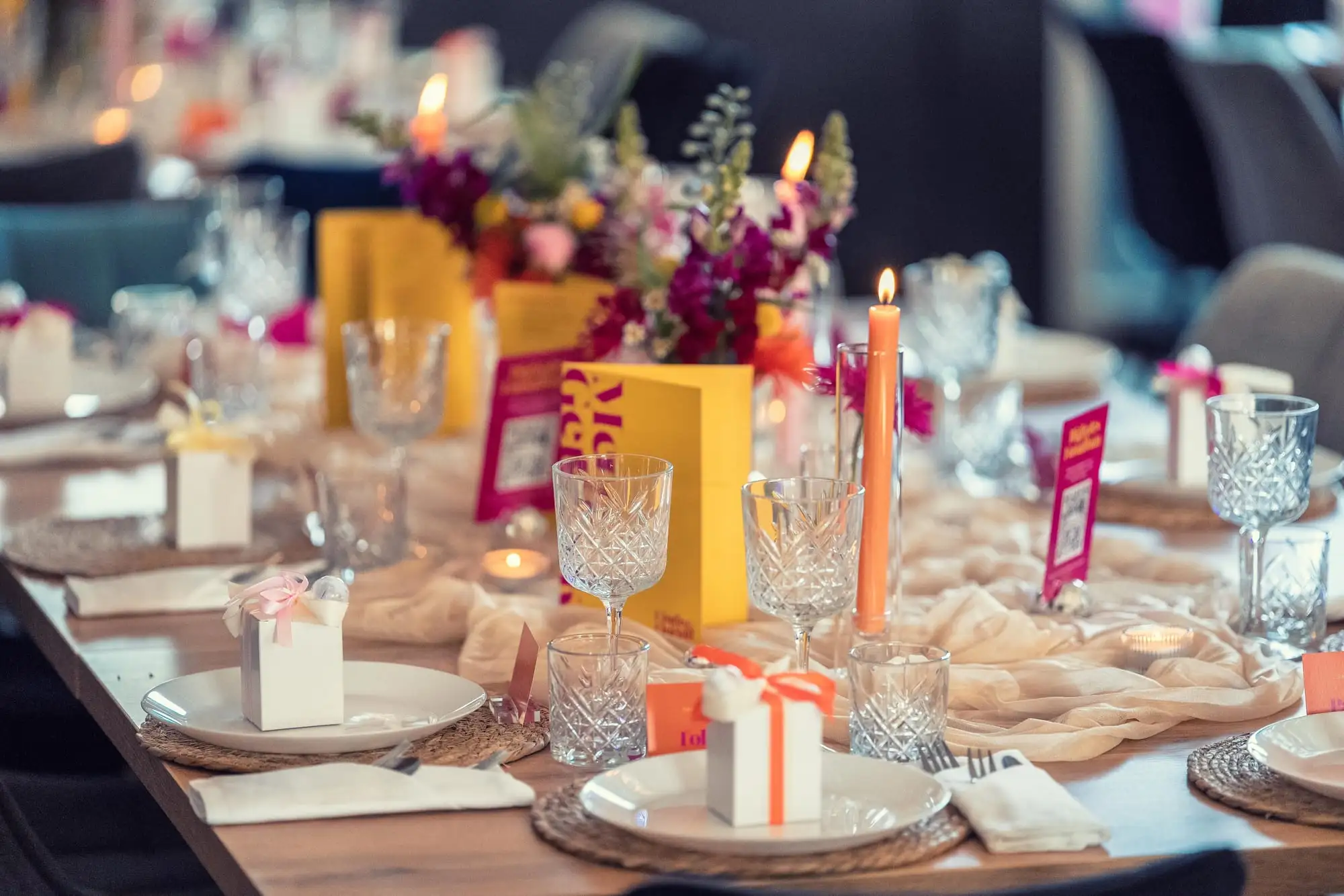 Gedeck mit Kristallgläsern, oranger Kerze und weißem Gastgeschenk vor gelben Menükarten