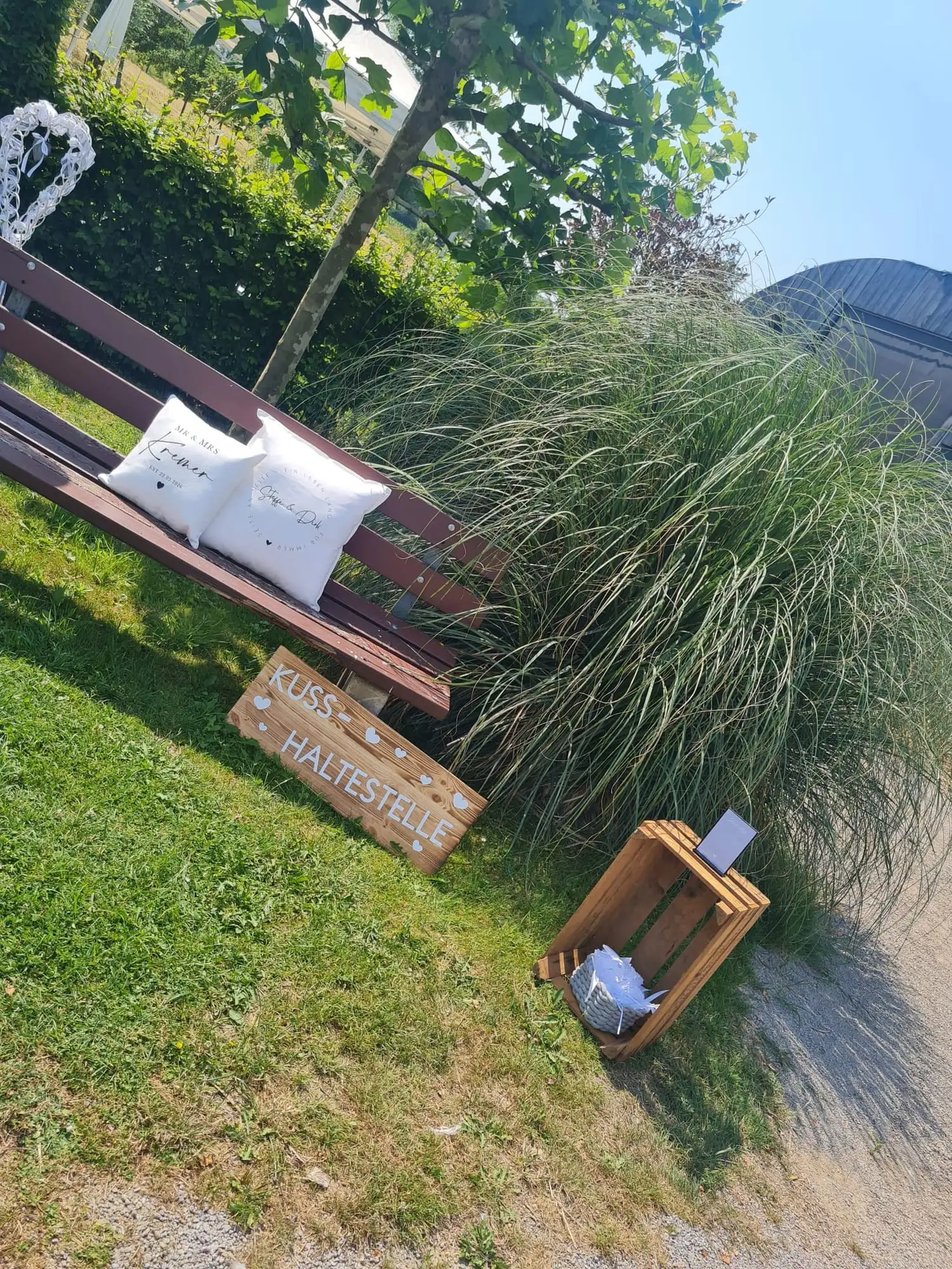 Holzbank mit bestickten Kissen neben Pampasgras, davor ein Schild mit der Aufschrift „Kuss-Haltestelle“