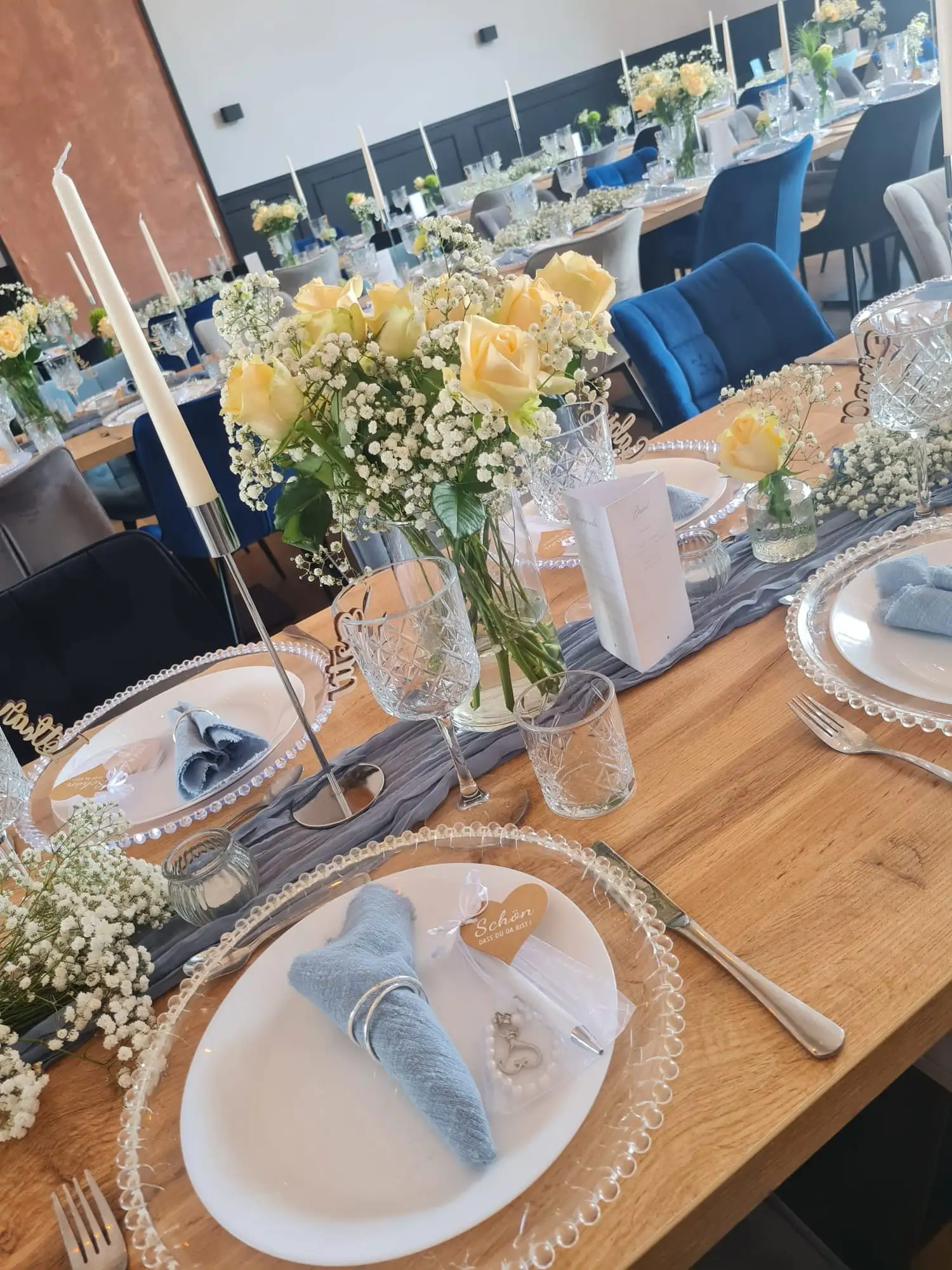 Gedeck mit gelben Rosen, Schleierkraut und blau gefalteten Servietten auf der Holztafel