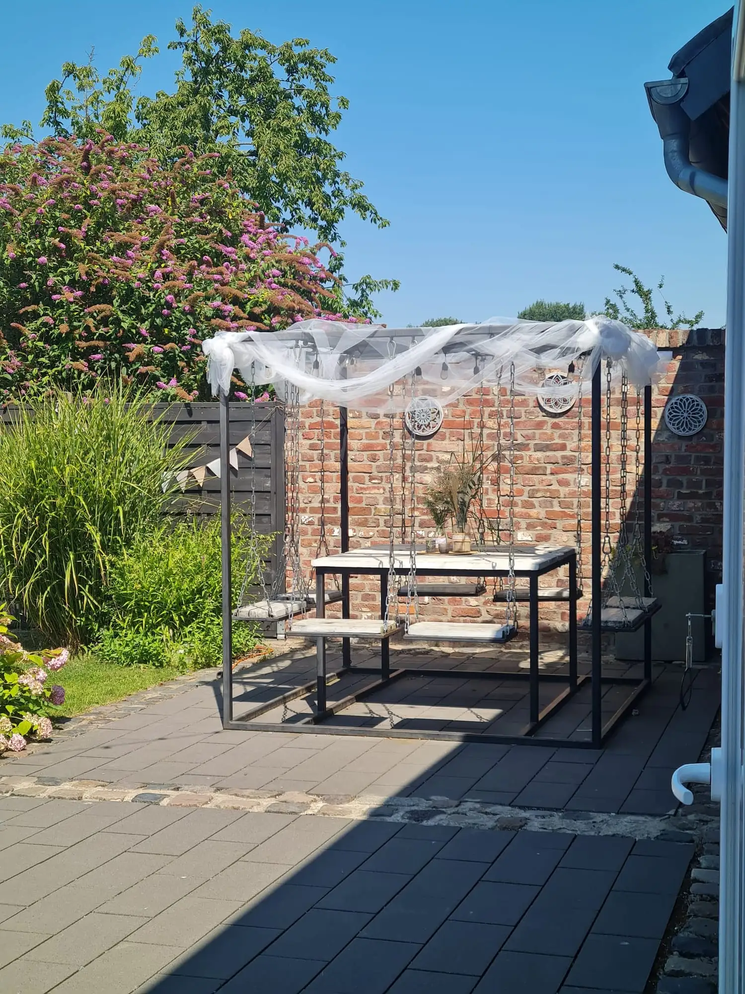 Tisch und Bänke unter einem schwarzen Gestell mit weißem Tuch an der Backsteinwand der Terrasse